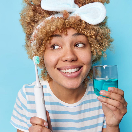 curly-haired-woman-brushes-teeth-indoor-holds-toothbrush-glass-mouthwash-takes-care-her-health-performing-daily-routine-wears-headband-casual-striped-t-shirt-isolated-blue-background.jpg